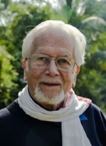 Das bild zeigt einen Mann mit weißen Haaren und Bart. Er hat eine Brille, ein blaues Hemd und einen weißen Schaal.